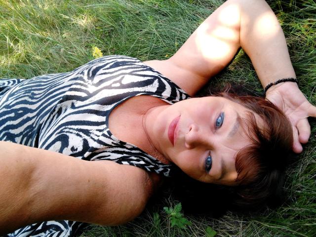MilaMils is lying on grass with zebra-patterned dress, arms above head, brown hair, blue eyes, and a black bracelet, looking at the camera with a relaxed expression.