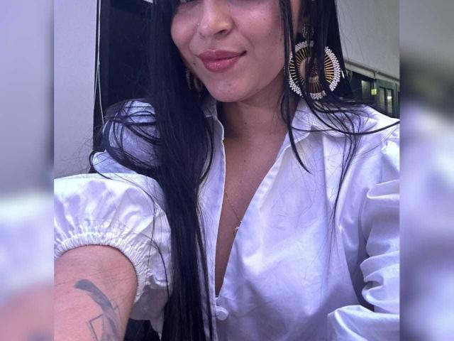 HikariChloe with long black hair, wearing a white button-up shirt, gold hoop earrings, and a subtle smile, showing a tattoo on her left arm, and a small cleavage.