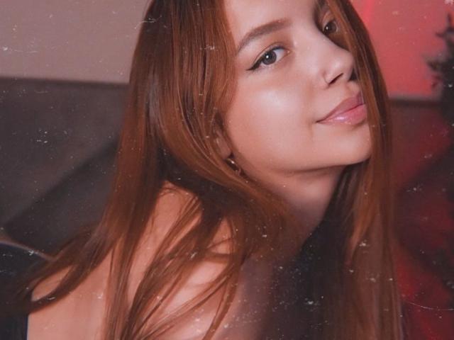 AstridWoods with long brown hair, soft smile, and subtle makeup, looking over her shoulder, showing her bare shoulders and a slight hint of her tits in the photo.