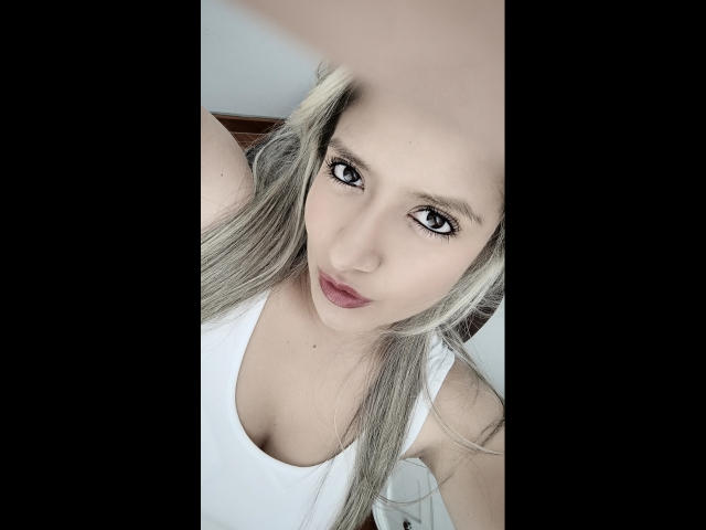 ValeriaHunter69 has blonde hair, blue eyes, and full lips. She's wearing a white tank top, showing her small, perky tits. She's looking up at the camera.