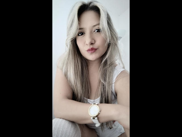 ValeriaHunter69 with blonde hair, white tank top, and gold wristwatch, sitting, looking at camera, slight pout, casual, sexy, attractive, soft lighting, white background, knee visible.