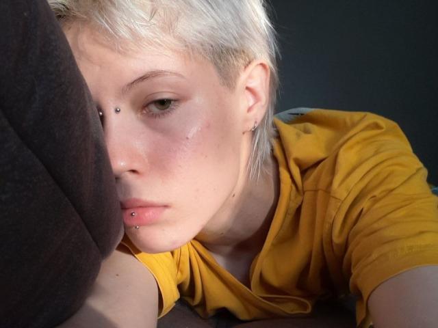MonyBabe has short, blonde hair, piercings on her nose and lower lip, and is wearing a yellow shirt, resting her head against a dark object.