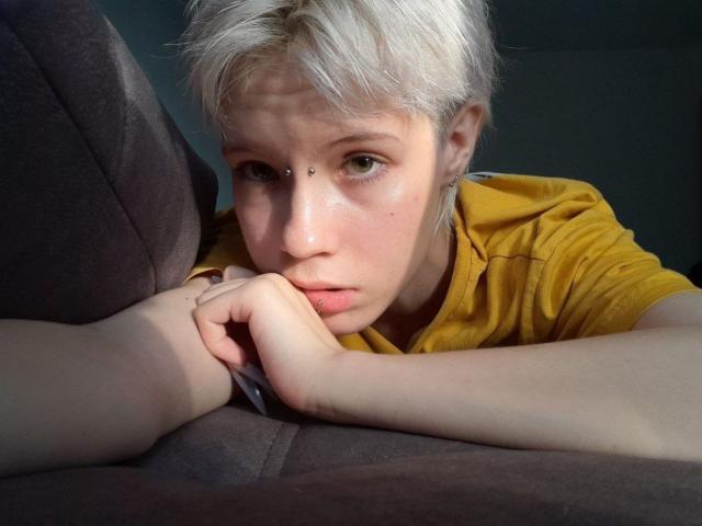 MonyBabe with short, white hair and a yellow shirt, resting her chin on her hand, looking tired with a small beauty mark above her nose.