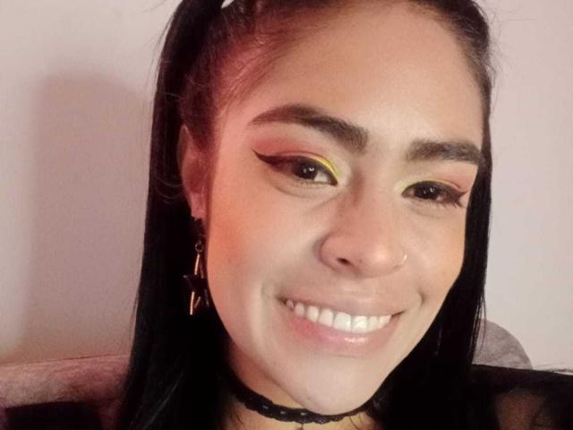 StellaBeckker with black hair in pigtails, yellow eyeliner, and a black choker, smiling with white teeth, nose ring, and gold earrings, in a selfie.