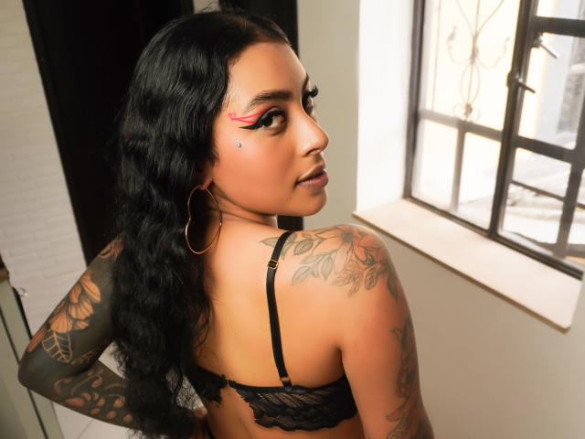 MagicMargo with long black wavy hair, red eyeliner, gold hoop earrings, black lace bra, tattooed arms and back, looking over shoulder, standing near a window.