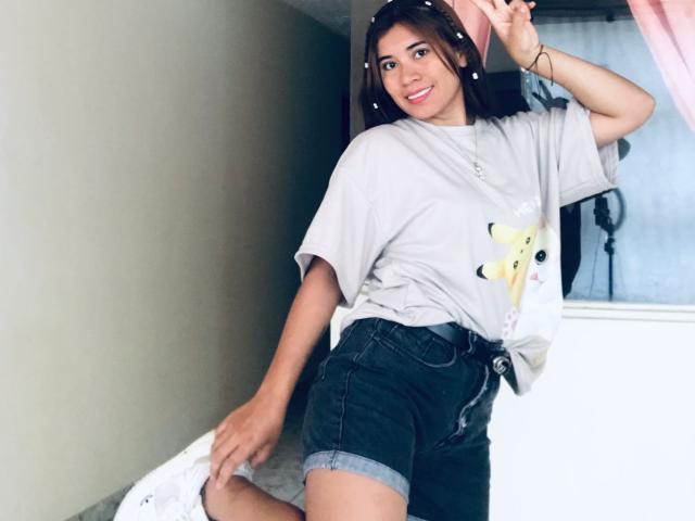 BellaBickers with brown hair, smiling, wearing a loose grey shirt, black denim shorts, and a hairband, showing a peace sign, standing with a white pillow in the background.