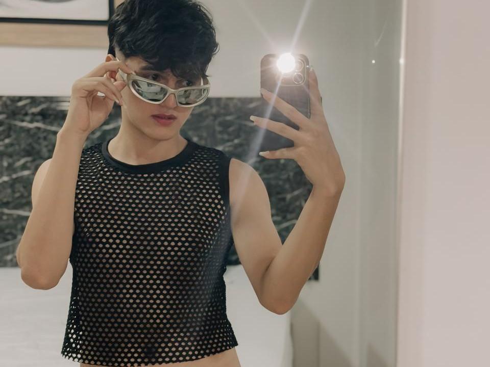 JacobI with short black hair, wearing a black mesh sleeveless top, glasses, and holding a phone in a mirror selfie, showing a flirty, confident expression and a small butt.