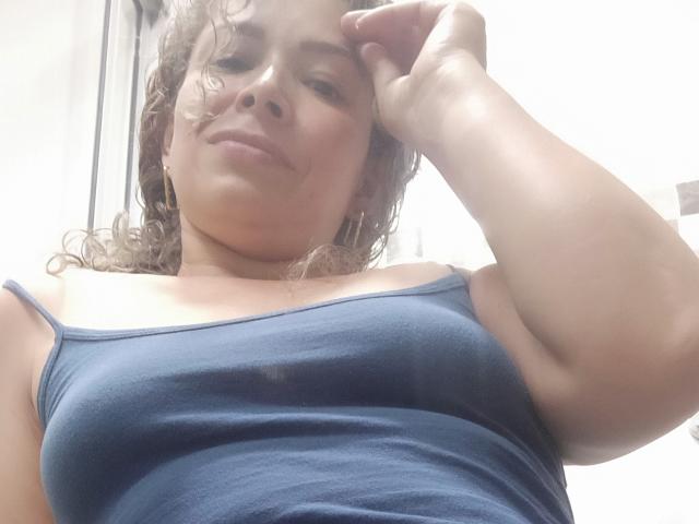 DalilaSuperHot with curly brown hair, blue tank top, showing her tits, slightly raised arm, confident smile, soft light, casual selfie, white background.