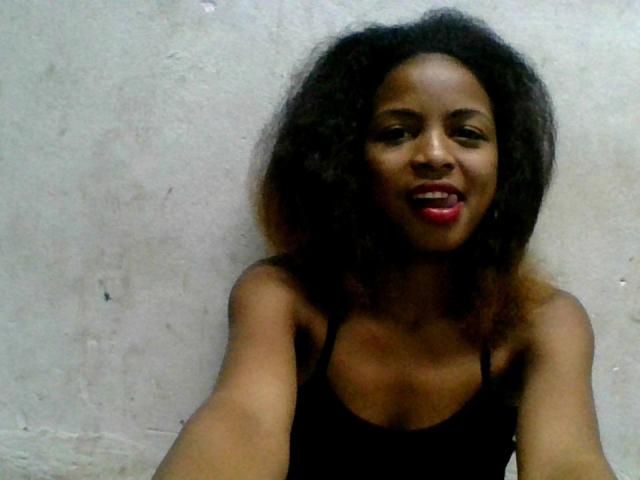 AlexaChan with dark curly hair, wearing a black tank top, smiling with red lipstick, sticking out her tongue, holding up a selfie. Her small tits are visible.