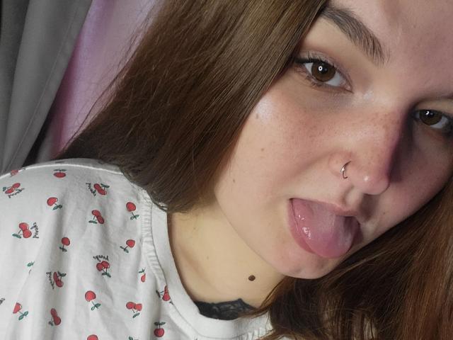 EvaBerg with brown eyes, nose ring, and freckles, tongue out, wearing a white shirt with red rose patterns, has a small mole under her chin.