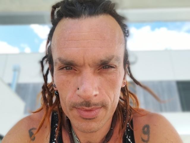 Chiroco tiene una frente arrugada, rastas marrones, un piercing en la nariz, tatuajes en los hombros y un rostro ligeramente fruncido con una expresión seria. Su camisa es negra y brillante..