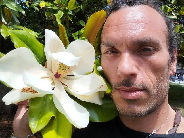 Chiroco with a large white flower and green leaves, holding it near his face, has a short beard, brown curly hair, and a slight smile, wearing a black shirt.