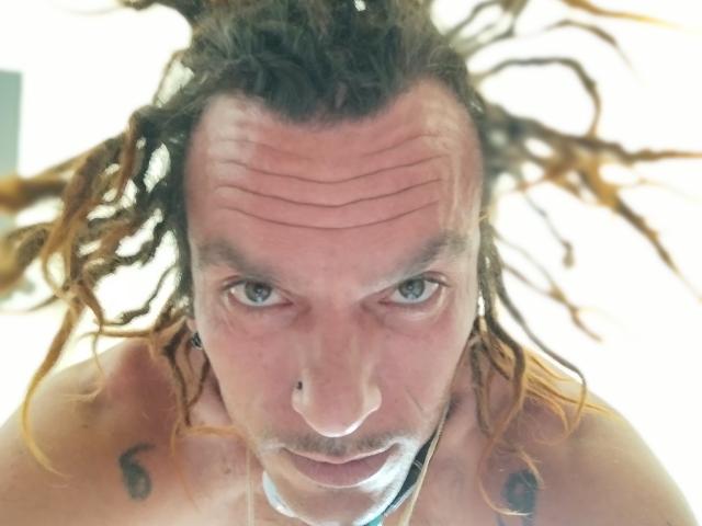 Chiroco tiene largas trenzas marrones, un piercing en la nariz y un tatuaje en el hombro derecho. Está mirando fijamente a la cámara con ojos azules, sin camisa..