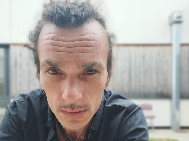 El rostro de Chiroco está centrado, con cabello oscuro y rizado y una expresión seria. Lleva una camisa negra, y sus ojos marrones miran directamente a la cámara..