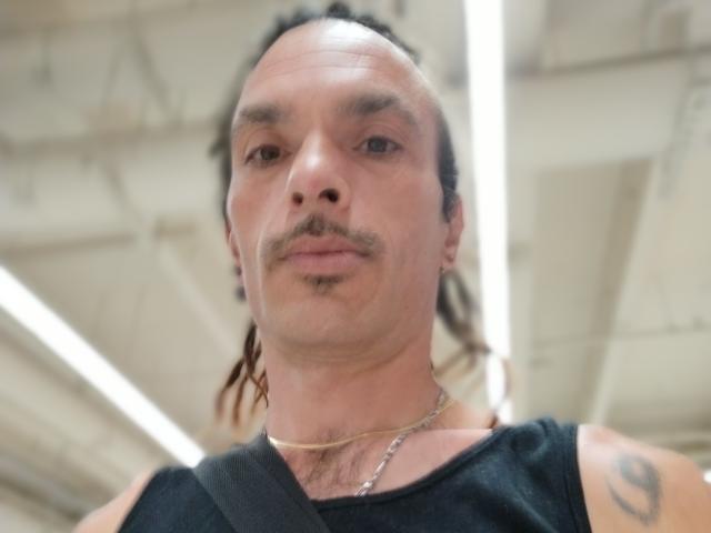 Chiroco has a balding head with long, dark, tied-back hair, a thin mustache, and a tattoo on his right arm. He's wearing a black sleeveless shirt and a necklace.