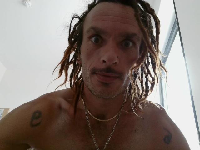Chiroco tiene un físico musculoso y sin camisa, con rastas, un bigote y tatuajes en ambos hombros. Lleva un collar de cadena y parece sorprendido..