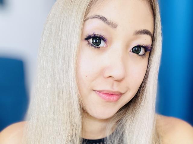 RenyLime has straight, platinum blonde hair, green eyes with purple eyeliner, and a small beauty mark on her left cheek. She's smiling with pink lipstick.