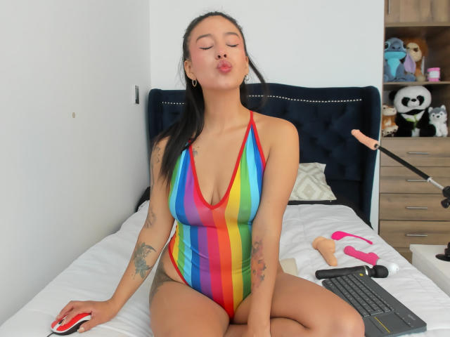 Soranaa with puffy lips, wearing a rainbow-striped one-piece, sitting on a bed, holding a red mouse, tattoos on arms, black hair, laptop, dildo, and vibrator in the background.