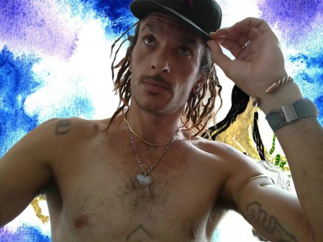Chiroco mit feuchten, braunen Dreadlocks, der eine schwarze Mütze trägt, ohne Hemd, mit Tattoos, Brusthaaren und einer Kettenkette, der in die Kamera schaut und dabei leicht grinst..