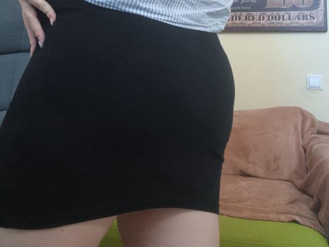 Leiliia's black skirt, tight against her ass, with a gray and white striped shirt, visible hand, and tan skin, standing in a room with a brown couch.