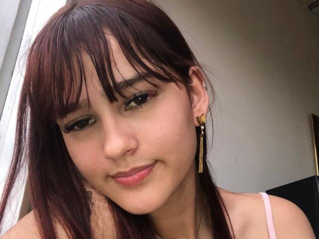 Melody Mansson a les cheveux bruns, avec une frange, et porte des boucles d'oreilles dorées avec des pièces rectangulaires. Elle sourit, avec du rouge à lèvres rose, des yeux marrons et un teint clair, et porte un haut rose..