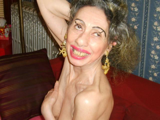 ElenahHottie, senza top, sorridente con rossetto rosa, orecchini dorati, braccio alzato, che tiene un seno, capelli ricci grigio-marrone, divano rosso, termosifone sullo sfondo, che appare felice e giocosa..