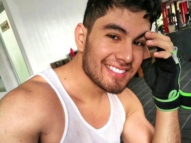 KevinAndres has a muscular build, short dark hair, and a beard, wearing a white tank top and black green gloves, smiling at the camera with a nose ring.