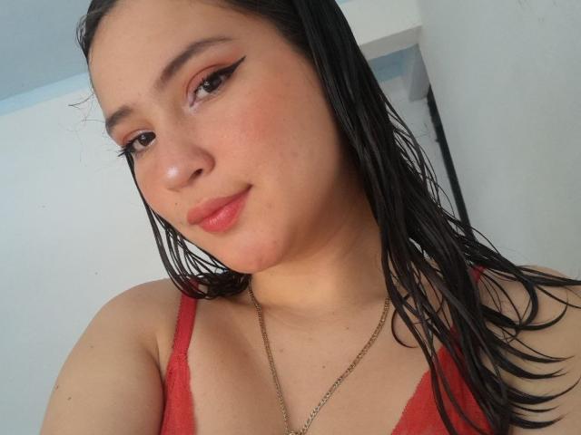 CelesteDash with wet black hair, red lipstick, and winged eyeliner, wearing a red tank top, showing her tits, and a gold necklace, smiling softly at the camera.