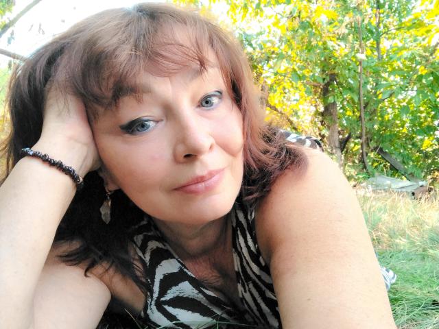 MilaMils with brown hair, blue eyes, and a black-and-white zebra-pattern dress, resting her head on her hand, smiling softly. Her tits are small, and she's outdoors.