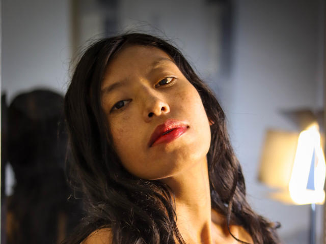 KarolineS has medium brown skin, black wavy hair, and red lips, looking slightly up with a relaxed, slightly seductive expression. Her small nose and subtle freckles are visible.