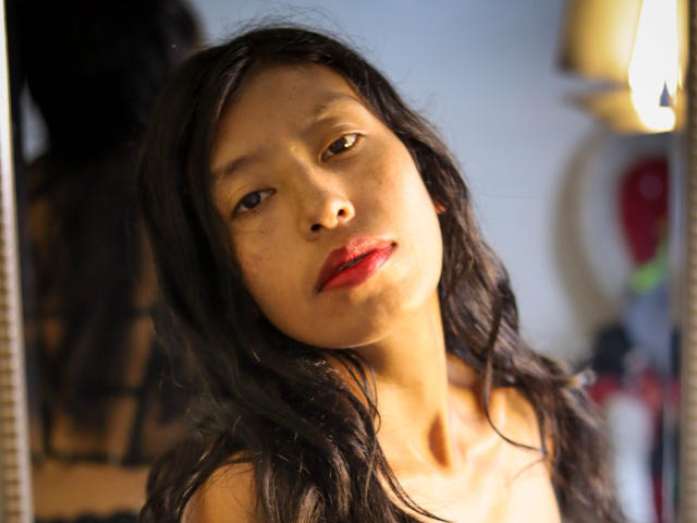KarolineS with long black hair, brown eyes, and red lipstick, slightly leaning forward, looking into a mirror, illuminated by a warm light, showing a soft, relaxed expression.