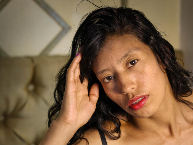 KarolineS has dark, wavy hair, brown eyes, and red lipstick. She's touching her head with a hand, showing a slightly pouty expression. Her tits are small and her pussy is not visible.