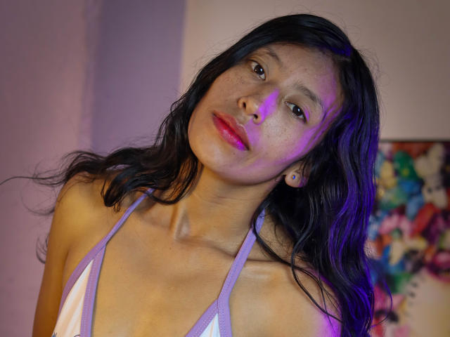 KarolineS with dark wavy hair, purple highlight on cheek, pink lipstick, and a white and purple halter top, looking slightly to the side with a subtle smile.