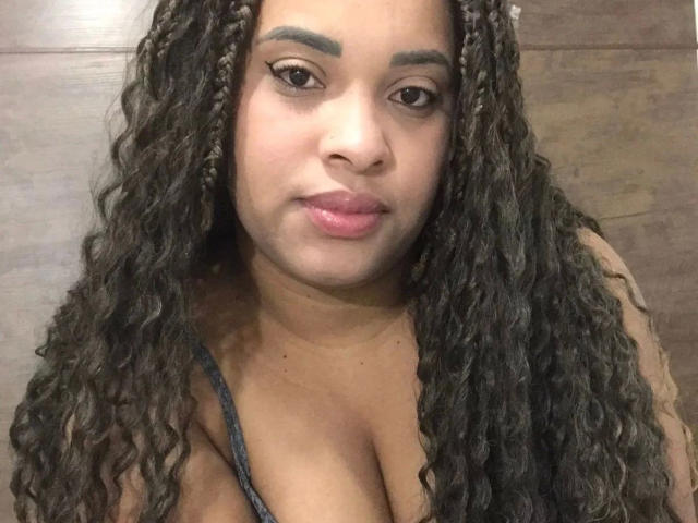 SuelenBarbara-hot has long curly brown hair, brown eyes, and full pink lips. She's wearing a gray top with visible cleavage. Her eyebrows are thick and she's looking at the camera.