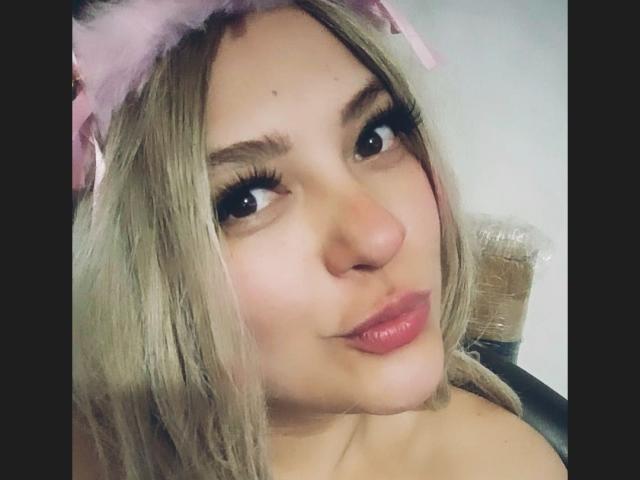 CorinaPai, con cabello rubio, una diadema rosa con plumas y labios rosados y fruncidos, mirando hacia arriba con pestañas largas y gruesas, ligeramente sonrojada, mostrando su linda nariz y labios carnosos y brillantes..