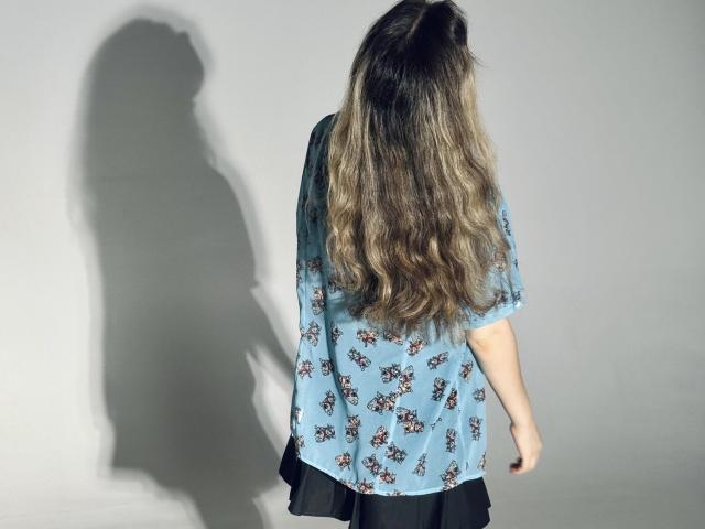 TinaRedly's long, wavy brown hair, wearing a loose blue floral shirt, shadow on gray wall, black skirt, back view, soft sunlight, subtle texture, casual style.