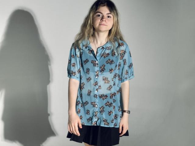 TinaRedly with long blonde hair, wearing a loose blue floral shirt, black pants, and a wristwatch, standing with a shadow on the wall.