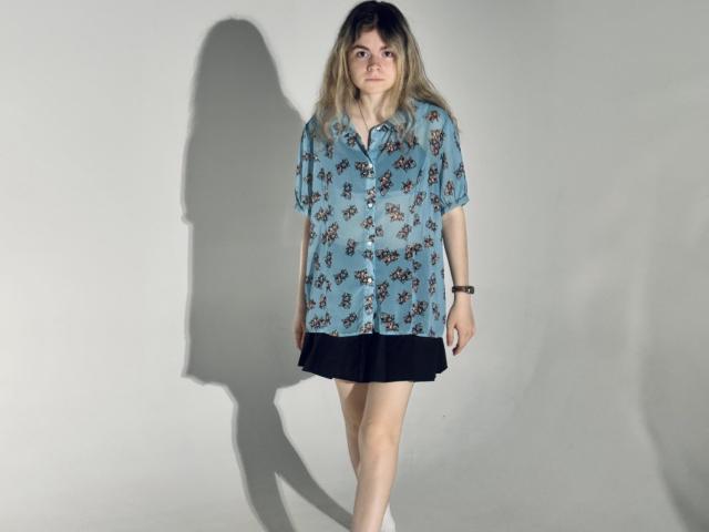 TinaRedly with light blonde hair, wearing a blue floral shirt, black skirt, and white socks, standing against a plain wall, casting a shadow, looking slightly up.