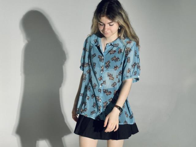 TinaRedly draagt een blauw bloesje met een zwarte rok, er is een schaduw op de muur, ze kijkt naar beneden, glimlacht, heeft blond haar, een polsbandje aan haar rechterpols, een casual stijl, zachte belichting..