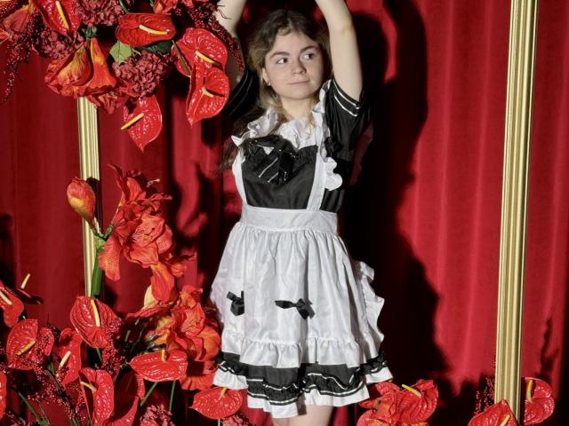 TinaRedly with curly brown hair, wearing a black and white French maid outfit, arms raised, standing in front of red flowers and a gold frame. She's looking to the side.