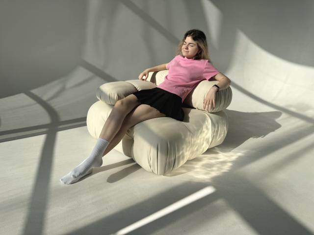 TinaRedly with pink top, black skirt, white socks, sitting on cream armchair, legs crossed, hands on arms, sunlight casting shadows, looking relaxed, blonde hair, subtle smile.