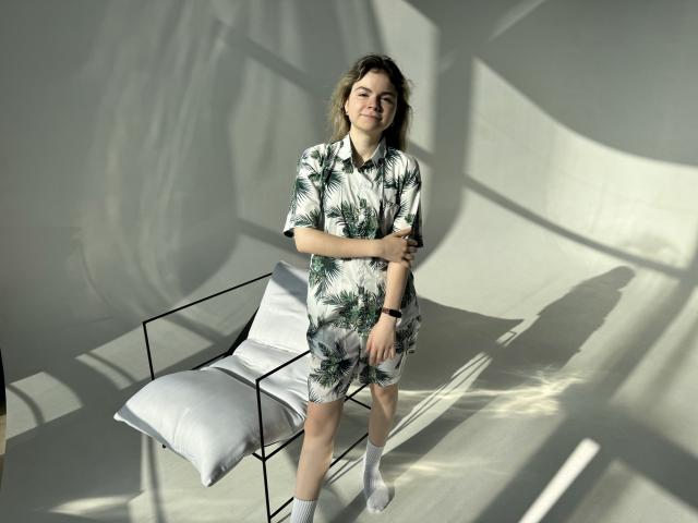 TinaRedly in a green leaf-patterned dress, standing with arms crossed, white socks, and black watch, smiling, next to a black chair with a white pillow.