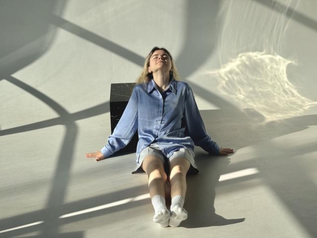 TinaRedly is sitting on a gray floor with arms out, wearing a blue shirt, shorts, and white socks, eyes closed, sunlight casting shadows, looking relaxed and content.