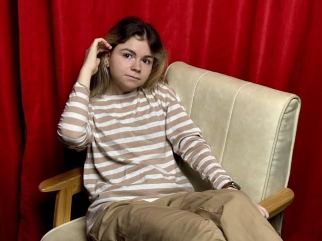 TinaRedly is sitting in a chair, wearing a striped brown and white shirt, beige pants, and wooden armrests, with red curtains behind her, touching her brown hair.