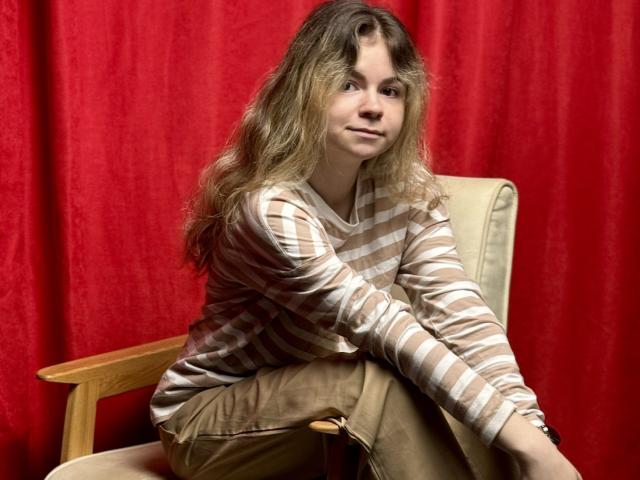 TinaRedly, with wavy blonde hair, sits in a wooden chair, wearing a striped shirt and brown pants, against a red curtain, looking slightly to the side.