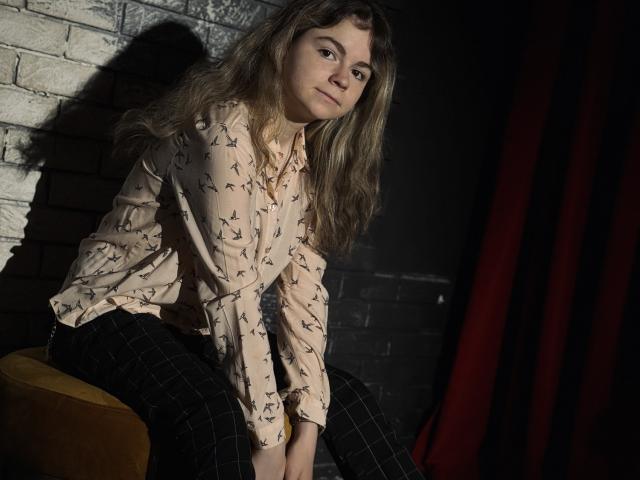 TinaRedly, sitting on a yellow stool, has long, wavy blonde hair, wearing a pink butterfly shirt and black checkered pants, with a serious, slightly annoyed expression.