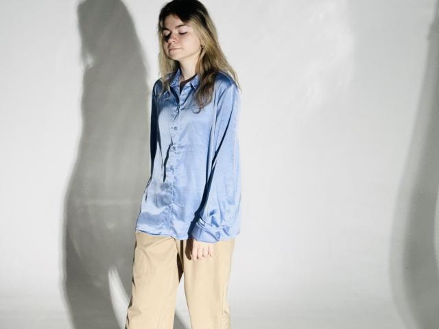 TinaRedly with long blonde hair, wearing a loose blue shirt and beige pants, standing, shadow on white wall, eyes closed, hands by sides, light from front.