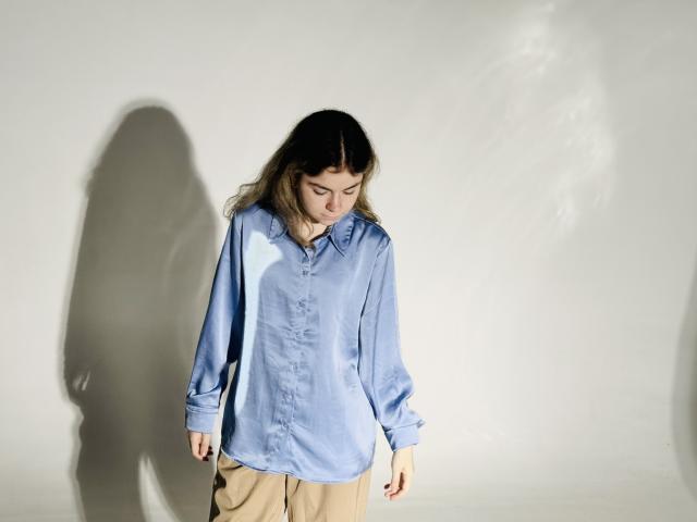 TinaRedly with long brown hair, wearing a loose blue shirt and beige pants, looking down, casting a shadow on a white wall. She's got a cute, shy vibe.