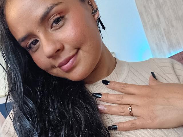 ArielleWilde with black hair, tan skin, and black nail polish, smiling softly. She's wearing a beige rib-knit top, a black ear ring, and a gold ring.
