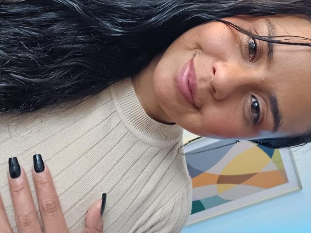 ArielleWilde with dark wavy hair, light brown skin, and black nails, wearing a beige ribbed sweater, smiling at the camera. Her hand rests on her chest.